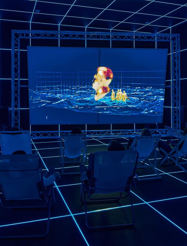 People seated in a dark room of a glowing blue grid, watching a surreal video.