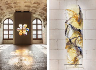 Partie gauche : Nous voyons une grande salle ancienne avec des murs et un plafond voûté en pierre sculptée. Deux grandes fenêtres à arc en plein cintre laissent entrer la lumière naturelle. Entre ces fenêtres, un mur moderne gris est installé, percé de huit cercles formant une fleur stylisée. Ces cercles sont constitués de vitraux colorés aux teintes chaudes (orange, jaune) et quelques touches de bleu et de violet, créant des motifs abstraits. La lumière traverse les vitraux et projette leurs couleurs vives