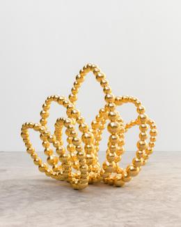 Une sculpture contemporaine composée de sphères dorées brillantes, reliées entre elles pour former des boucles sinueuses et aériennes. Chaque boucle rappelle la forme d'un pétale, créant ensemble une structure florale abstraite qui évoque un lotus stylisé. L’œuvre est posée sur un sol en béton clair, dans un espace minimaliste aux murs blancs, mettant en valeur l’éclat doré des sphères.