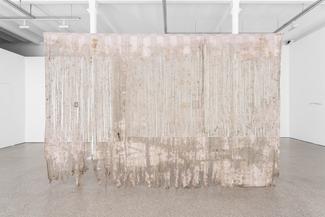 Grande pièce de tissu translucide suspendue, aux teintes pâles et abîmée en bas, avec des motifs verticaux blancs, dans une galerie blanche minimaliste