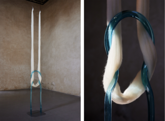 Une installation artistique contemporaine montrant une structure en verre bleu transparent en forme de boucle verticale. Deux longues mèches de fibres blanches (ressemblant à de la laine ou du coton) passent à travers la boucle et tombent jusqu'au sol. L’image de gauche montre l’installation entière dans une pièce aux murs en plâtre beige, tandis que l’image de droite offre un gros plan sur la texture des fibres et la transparence du verre.