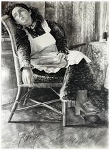 Assis, fauteuil rotin, robe chat tablier, regarde à droite - 1983