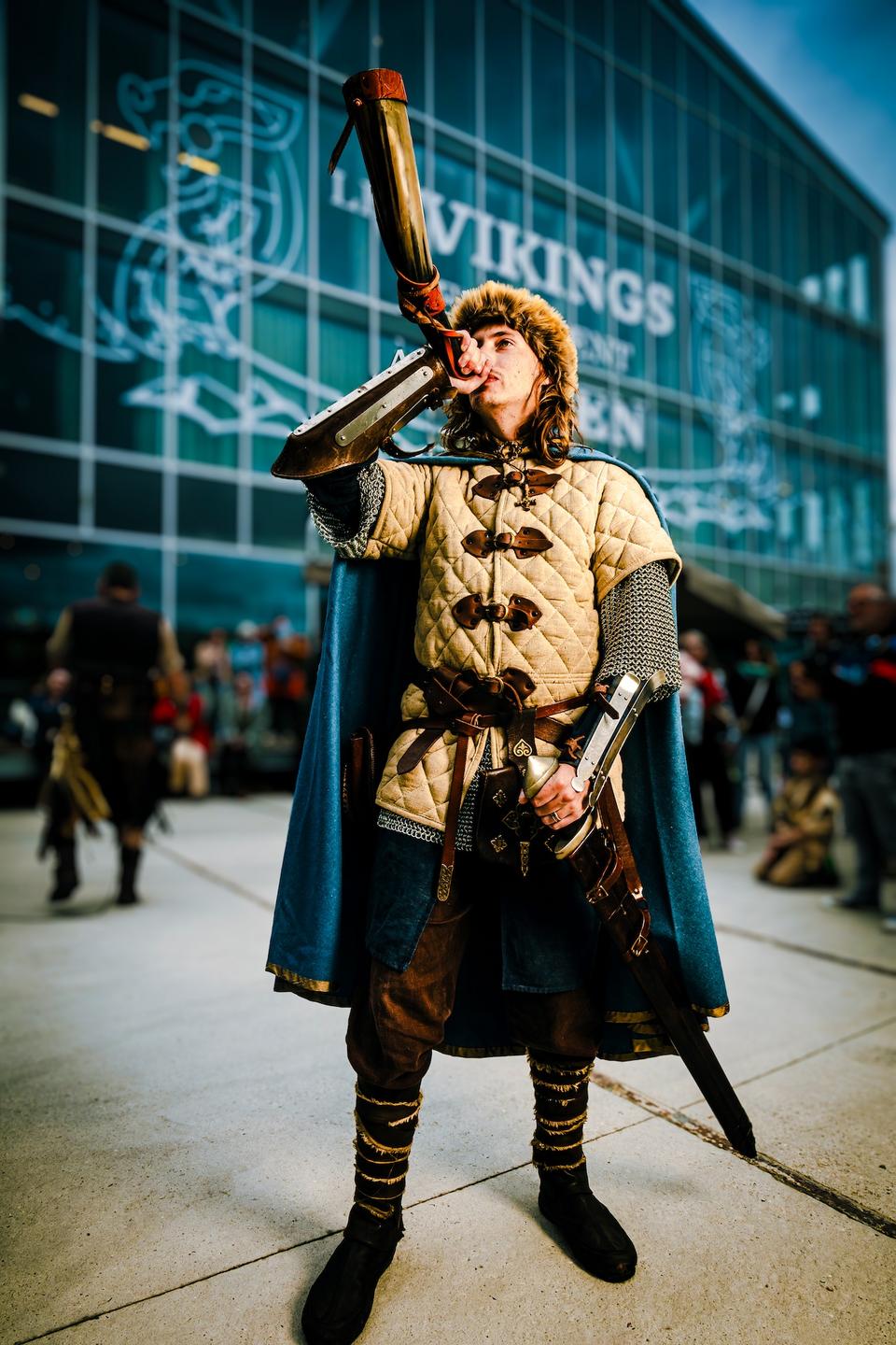 Homme en costume viking soufflant dans un cor devant un bâtiment vitré lors d’un événement festif.